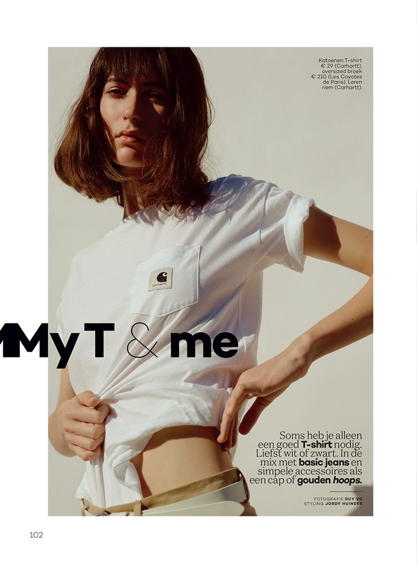 A young adult woman poses in a studio wearing a white Carhartt t-shirt and light-colored belt. She is looking at the camera.