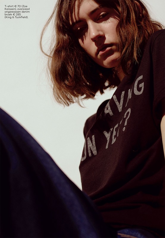Brunette woman poses in black t-shirt and blue jeans against a white background, captured in a fashion photography style with a low angle.