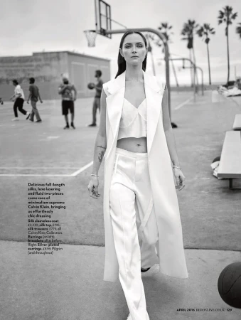 Monochrome fashion shot of a woman in a white silk outfit on a basketball court, surrounded by palm trees and blurry background figures.
