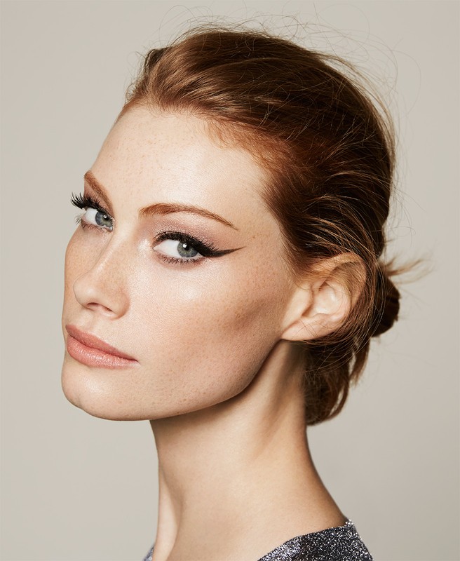 Close-up beauty portrait of a red-haired adult woman wearing makeup and a dark, shimmering top against a neutral background.
