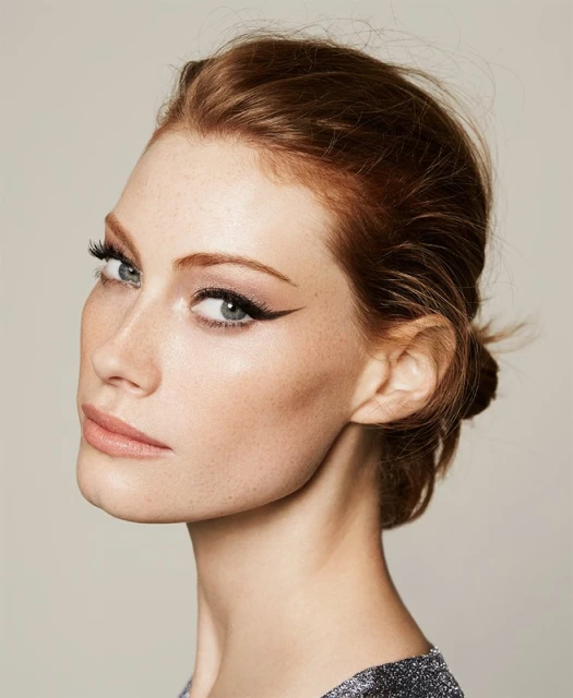 Close-up beauty portrait of a red-haired adult woman wearing makeup and a dark, shimmering top against a neutral background.