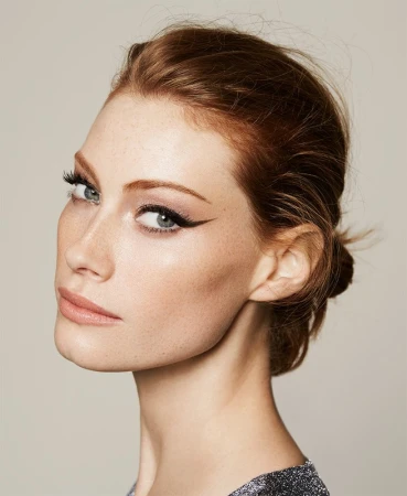 Close-up beauty portrait of a red-haired adult woman wearing makeup and a dark, shimmering top against a neutral background.