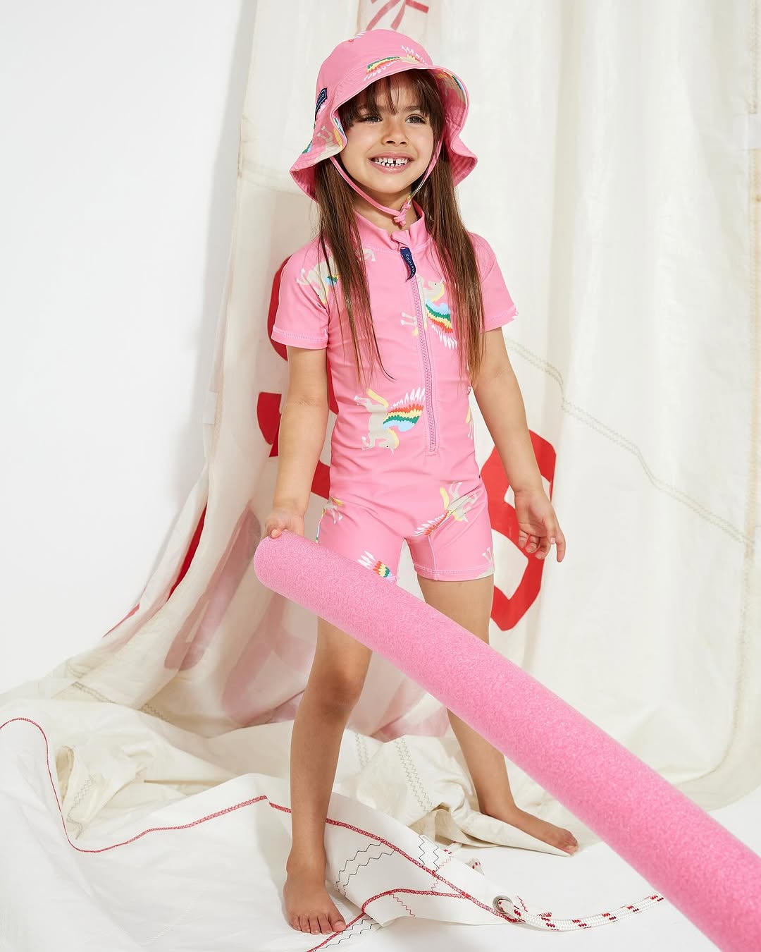 Young girl in a pink swimsuit and bucket hat poses with a pink foam pool noodle against a white background with nautical sail.
