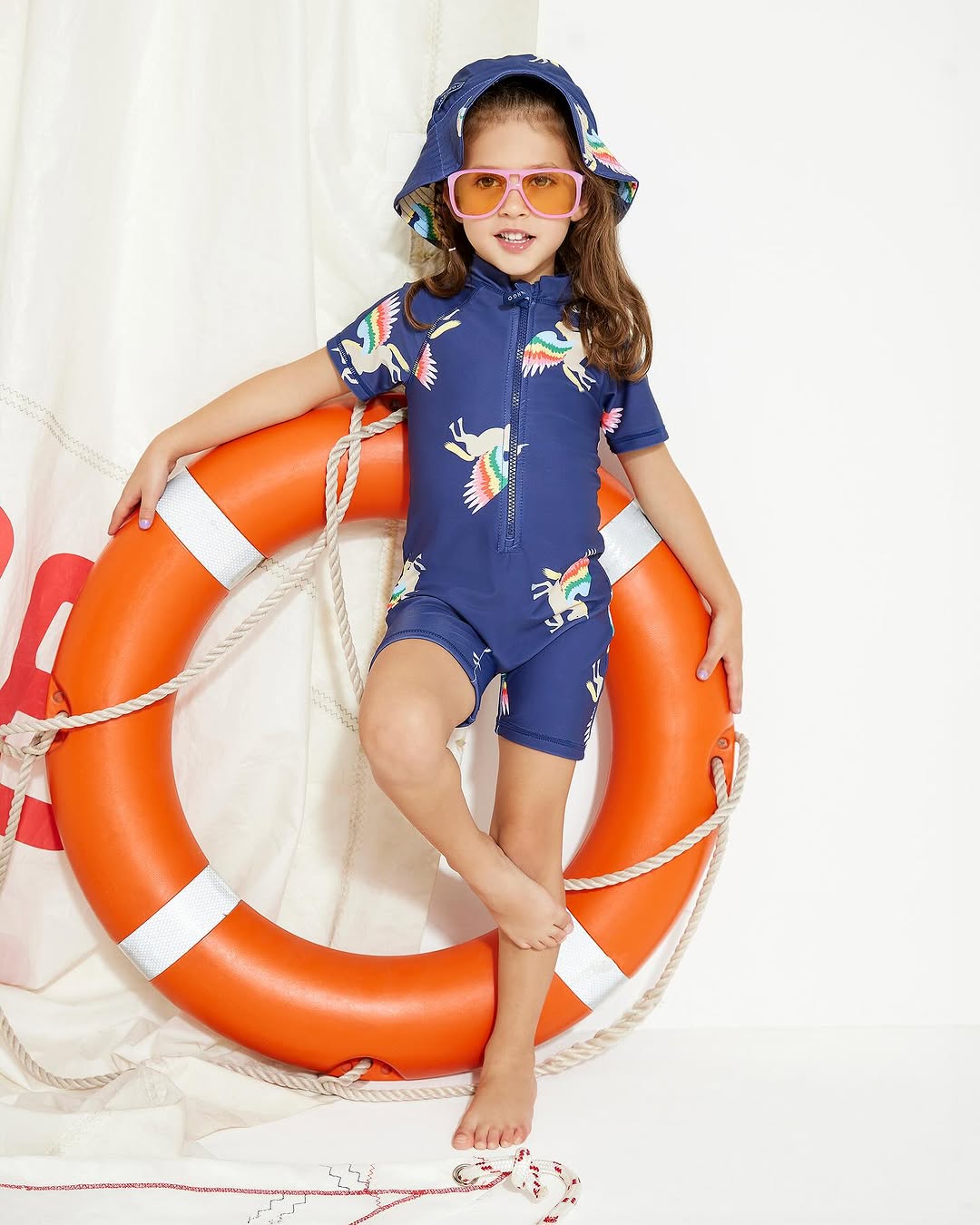 Child in a unicorn swimsuit and sun hat stands with a life preserver. The subject wears orange sunglasses against a white backdrop.