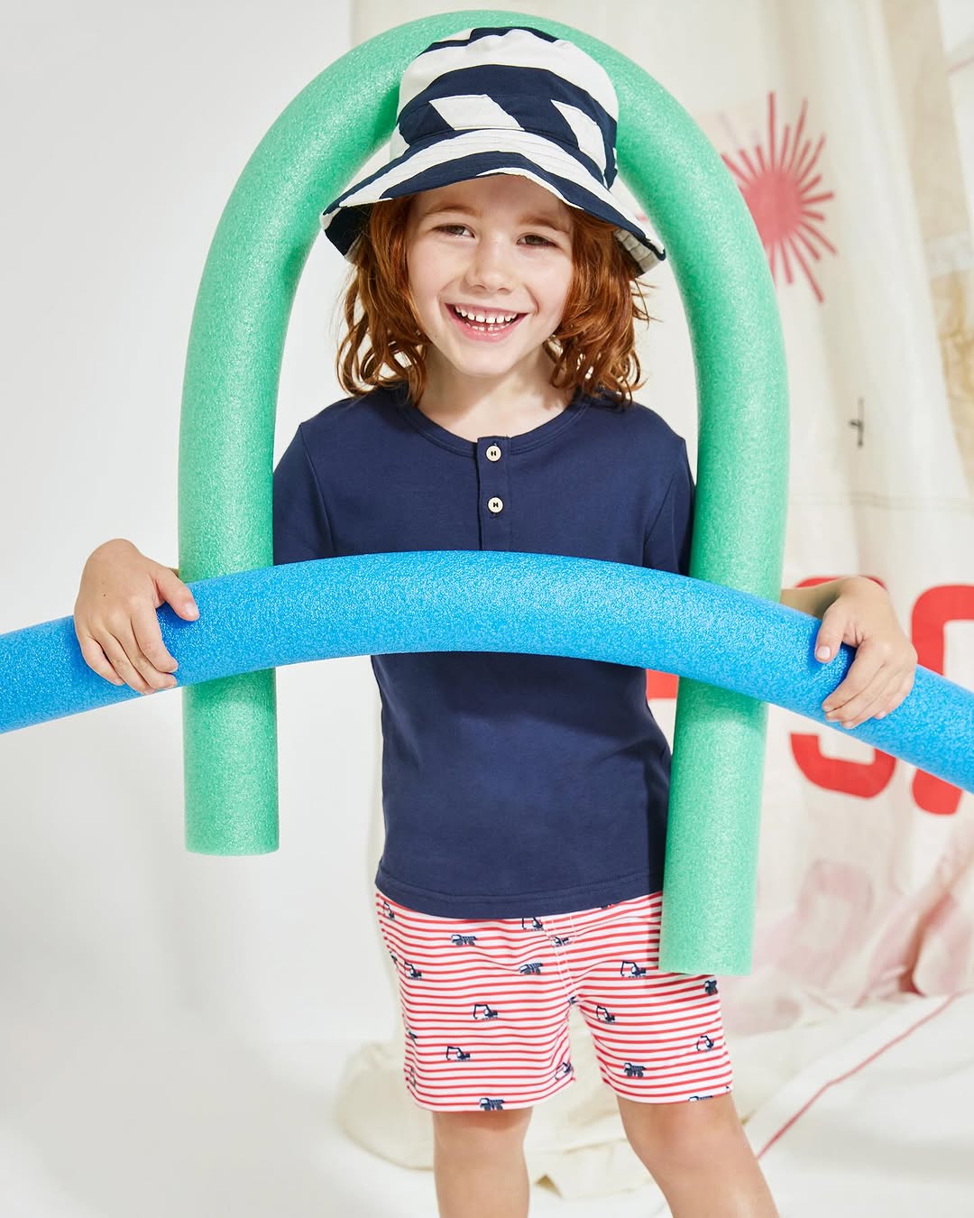 Smiling young child in summer clothing holds blue and green foam pool noodles in a brightly lit studio environment.