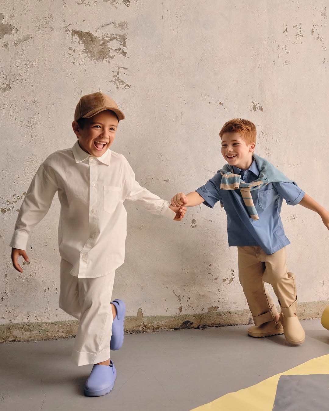 Two young children holding hands, laughing, and running in a bright indoor setting. One wears a hat and the other has a sweater around his neck.