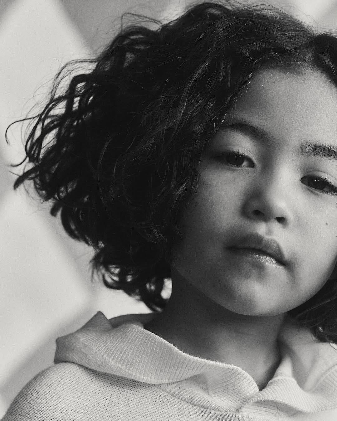 Portrait of a young Asian girl with curly dark hair wearing a light-colored hoodie, close-up shot in a softly lit indoor setting.