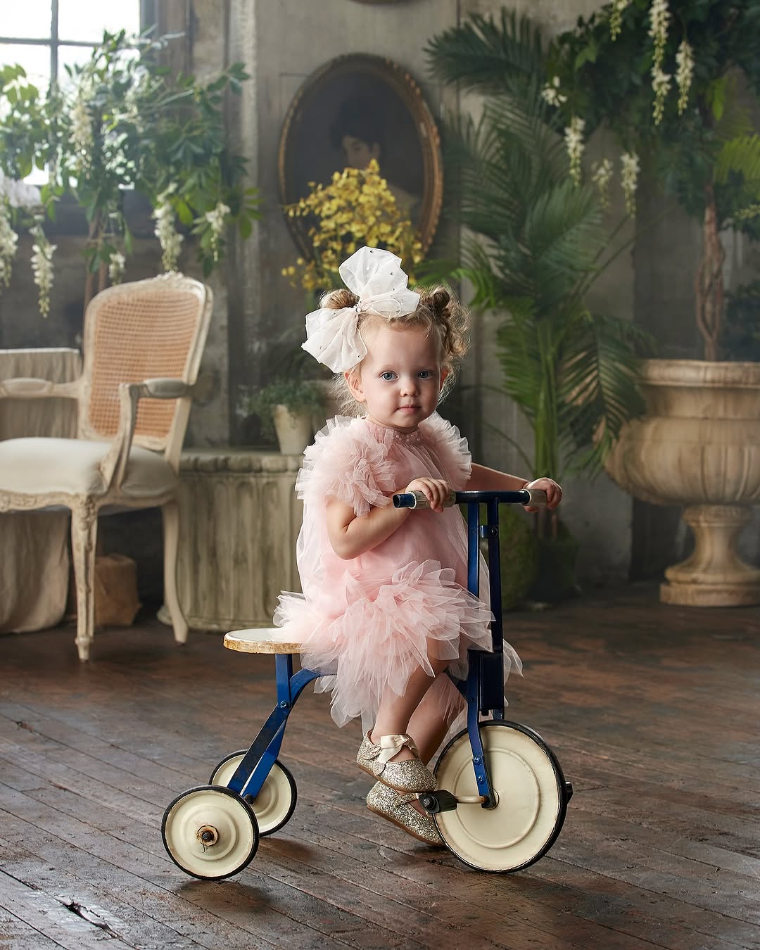 A young child rides a vintage tricycle indoors, wearing a pink dress and sparkly shoes, with decorative plants and furniture in the background.