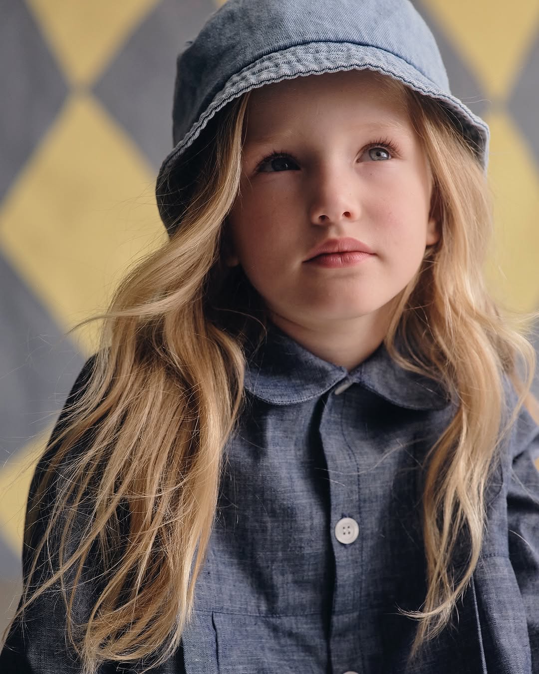Close-up of a young girl with long blonde hair, wearing a blue denim outfit and a matching hat, looking up in a soft indoor setting.
