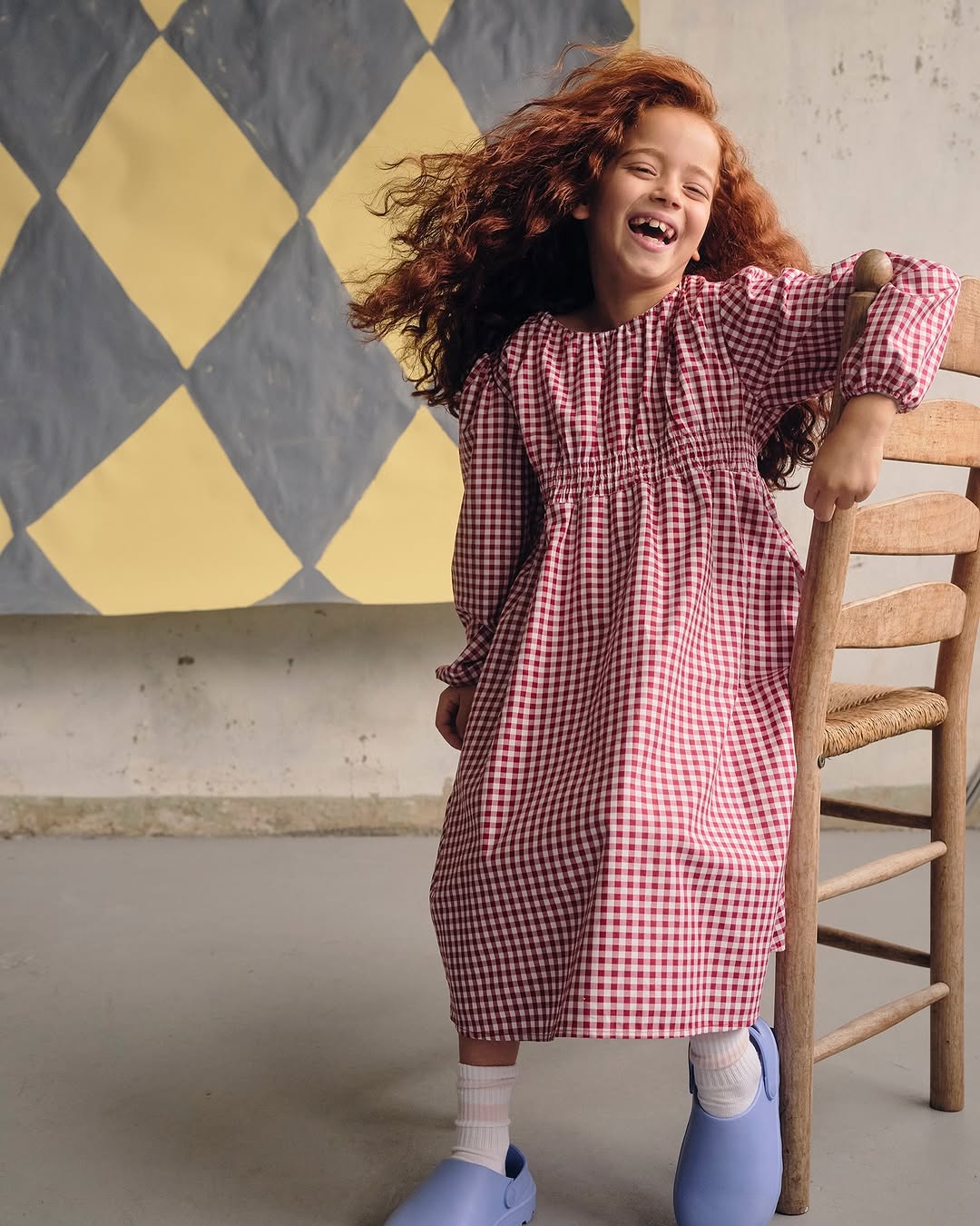 A young red-haired girl in a red-and-white gingham dress smiles indoors near a vintage chair and a diamond-patterned backdrop.
