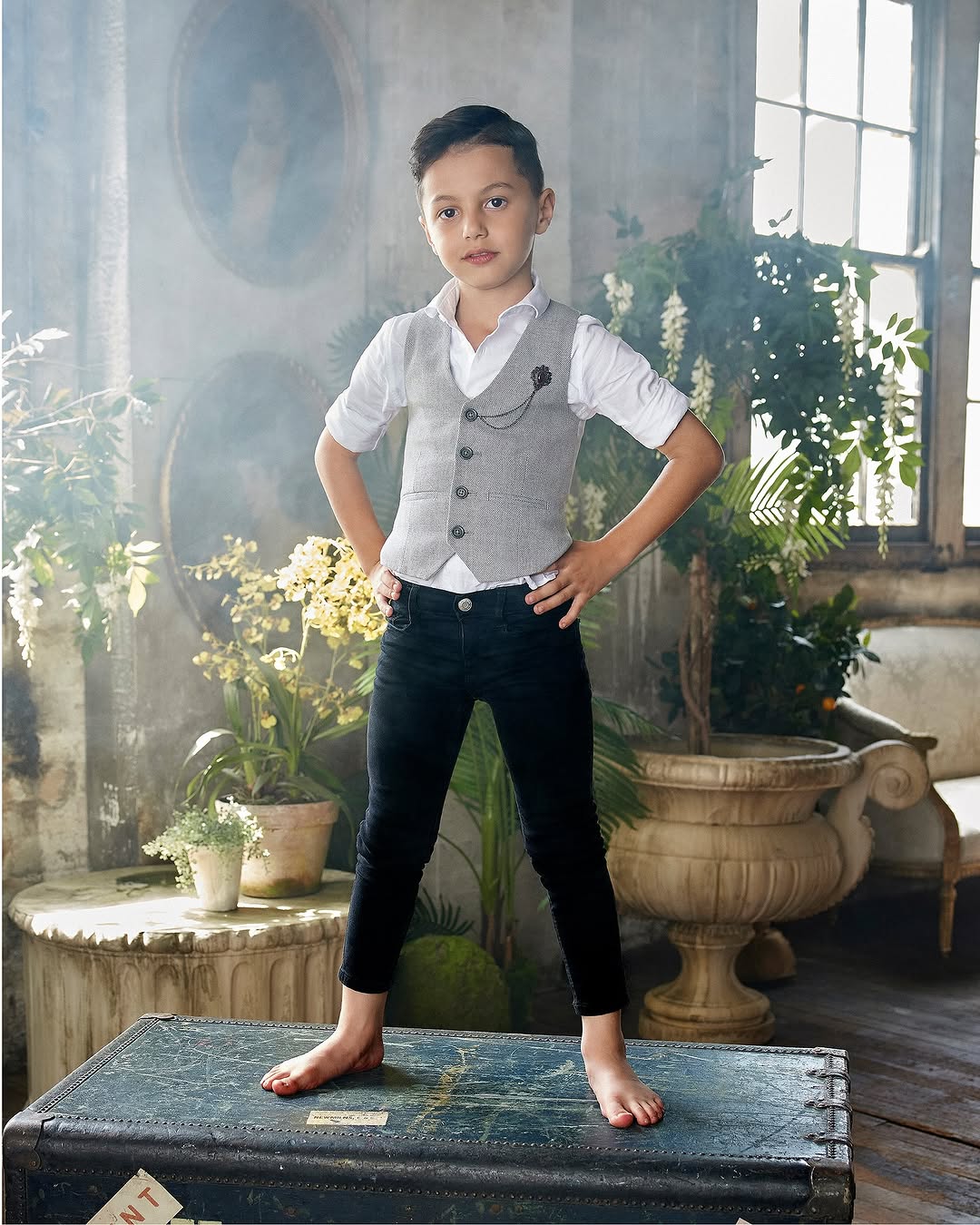 A stylish young boy poses barefoot on a vintage trunk, wearing a vest, white shirt, and black jeans in an indoor setting.