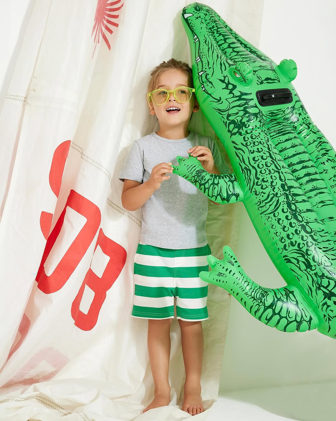 Smiling young boy wearing yellow glasses and striped shorts, posing next to a large inflatable green alligator toy against a white backdrop.