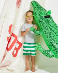 Young child wearing yellow glasses and green and white striped shorts holding an inflatable green alligator toy.