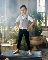 A young boy standing barefoot on a vintage trunk wearing a grey waistcoat, white shirt, and dark trousers.