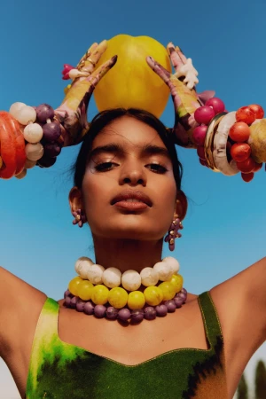 Model holding a bright yellow fruit above head, wearing stacked colourful bead necklaces and patterned gloves against clear blue sky.