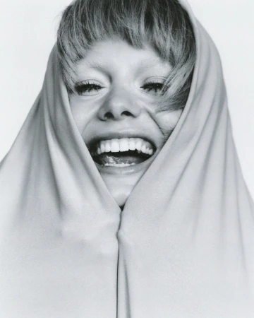 Close up portrait of a laughing woman with bangs framed by light fabric against a white seamless background.
