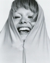 Close up portrait of a laughing woman with bangs framed by light fabric against a white seamless background.