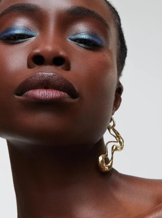 Close-up portrait of a Black woman with metallic blue eyeshadow and glossy lips wearing a sculptural gold earring