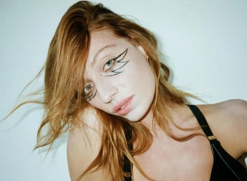 Close-up portrait of a woman with red hair and graphic winged eyeliner wearing a black top against a plain white wall