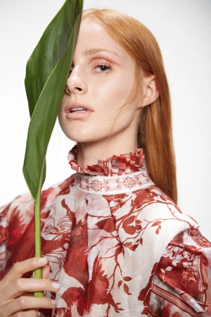 Red-haired female model holding a large green leaf across her face wearing a patterned red and white high neck dress against white.