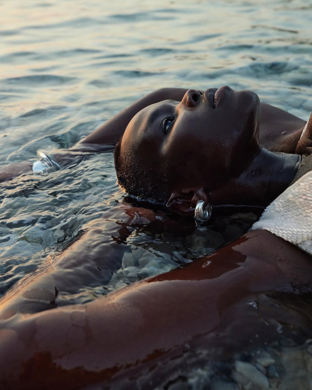 Black woman floats serenely in gentle ocean waves. Her arms are outstretched. She wears silver earrings and a light-colored cover-up.