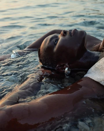 Black woman lying in shallow water with wet skin and hair, wearing silver hoop earrings and a white textured top.