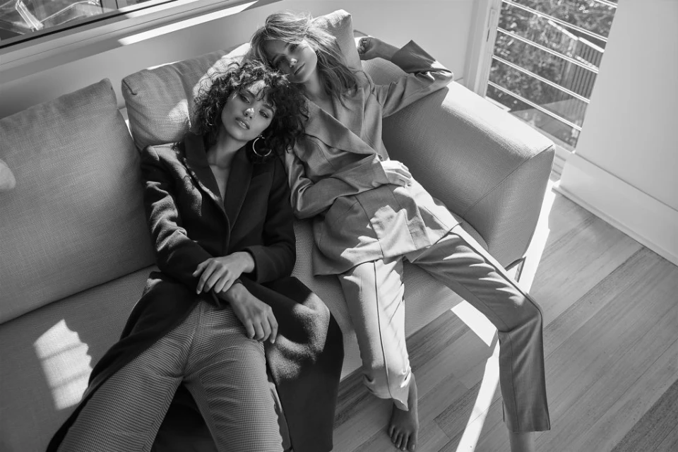 Two women in suits lounging on a couch in a brightly lit room, showcasing fashion and style in a relaxed setting, monochrome.