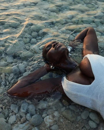 Black woman wearing a white swimsuit lying in shallow clear water over smooth round pebbles, photographed by Emily Abay.