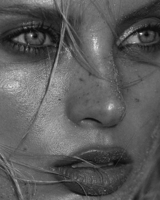 Close-up black and white portrait of a woman's face with glistening skin, prominent freckles, and strands of blonde hair.