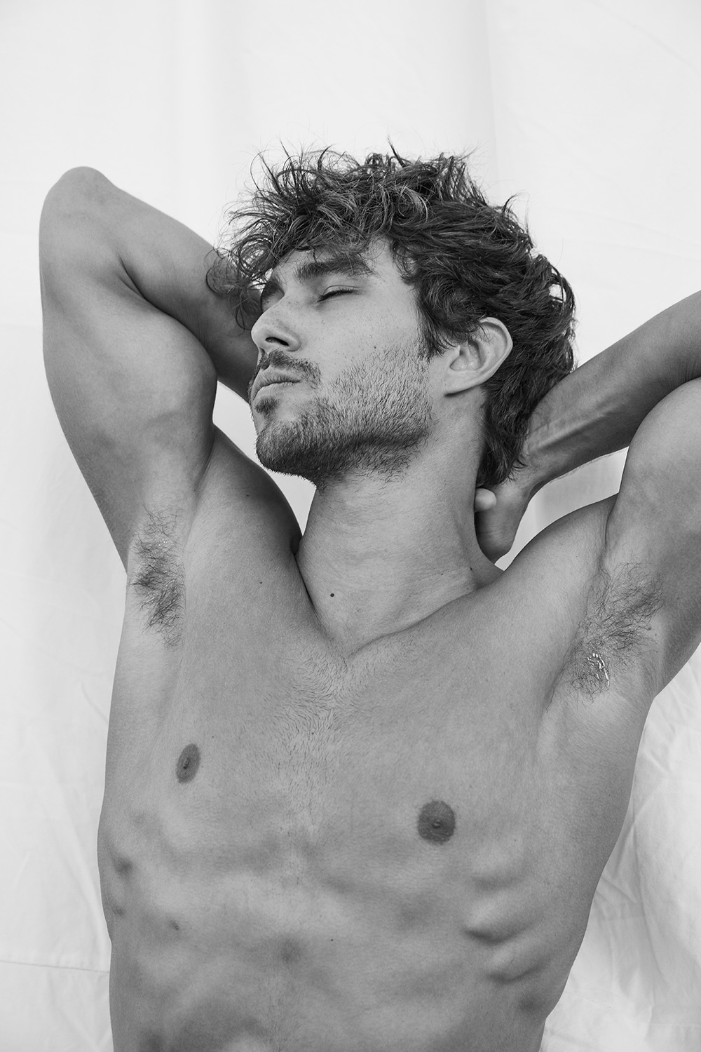 Black and white portrait of a shirtless male model with curly hair and closed eyes, arms raised behind his head against a white backdrop.