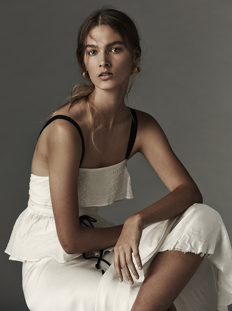 Elegant portrait of a young woman in a white dress with black straps, seated indoors against a grey backdrop with soft, diffused lighting.