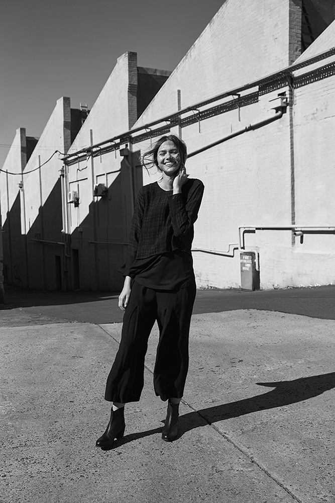 Black and white portrait of a smiling woman wearing black pants and a long-sleeved shirt, posing against a textured industrial building.