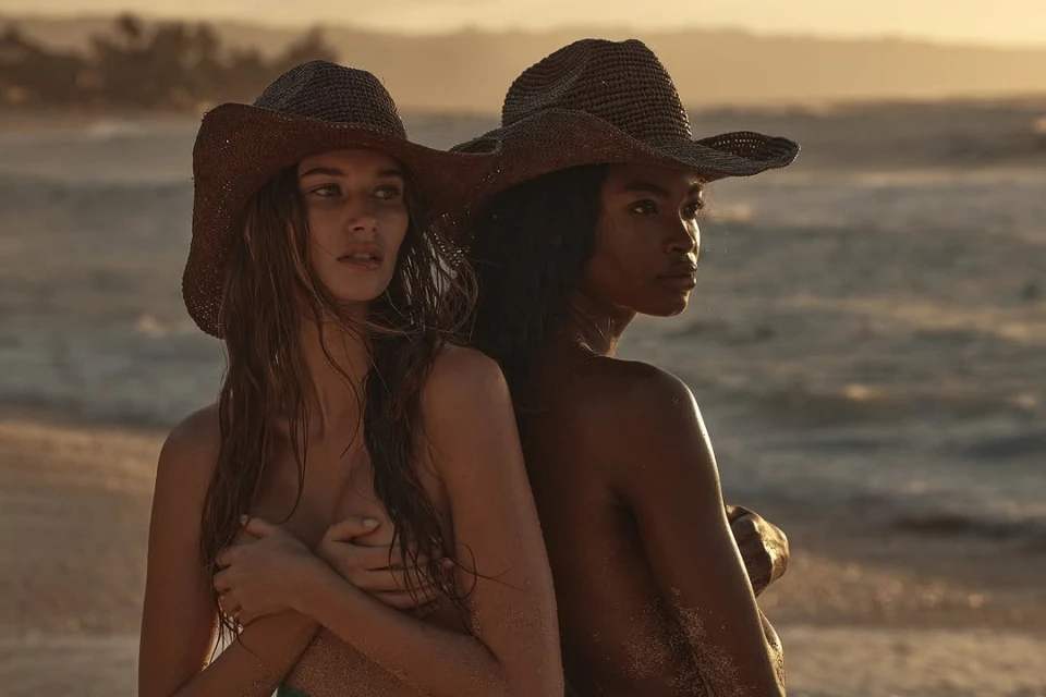 Two women with straw hats pose on a beach with a serene ocean backdrop at sunset. Fashion, travel and nature photography.