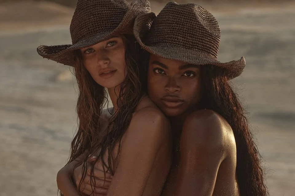 Two women embrace on a beach, wearing woven hats, with soft, warm lighting highlighting their skin tones and natural beauty.