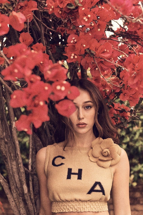 A young woman wearing a beige knit top with a large flower detail stands framed by vibrant red bougainvillea flowers.