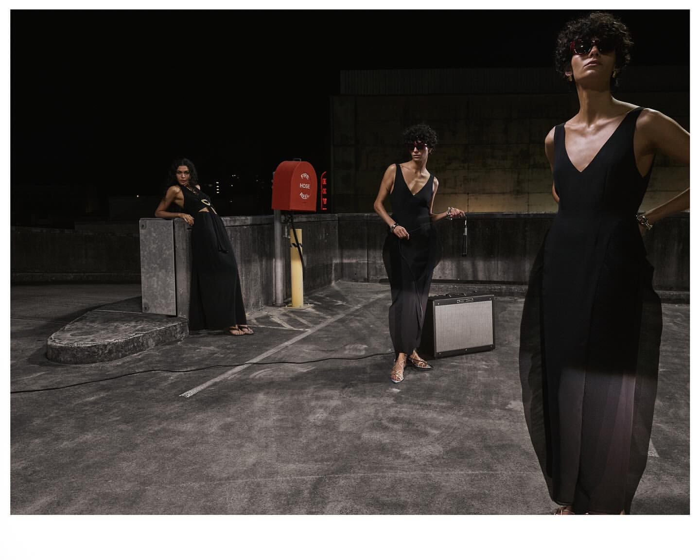 Three women in stylish black dresses pose on a rooftop at night, showcasing fashion with an urban backdrop, amplified by studio lighting.