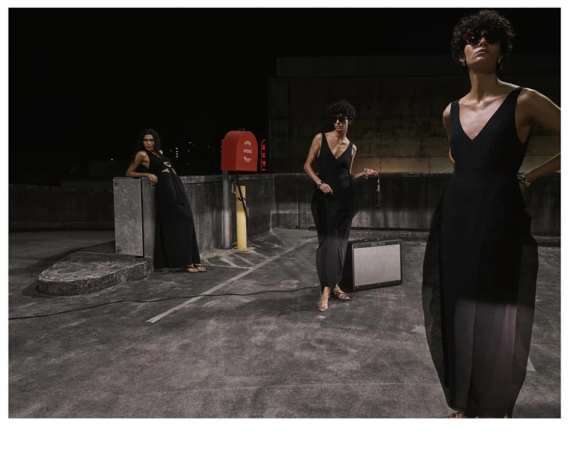 Three women in stylish black dresses pose on a rooftop at night, showcasing fashion with an urban backdrop, amplified by studio lighting.