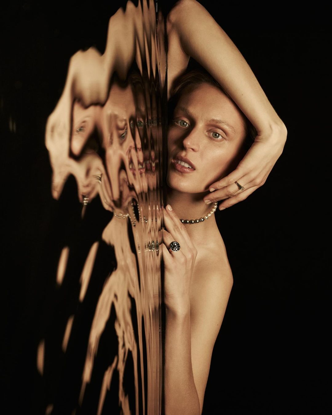 Portrait of an adult woman posing in front of a dark background, partially obscured by textured glass and wearing jewelry.