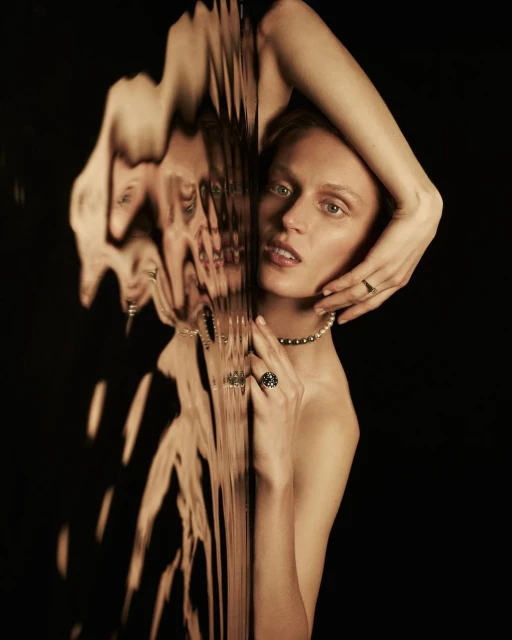 Portrait of an adult woman posing in front of a dark background, partially obscured by textured glass and wearing jewelry.