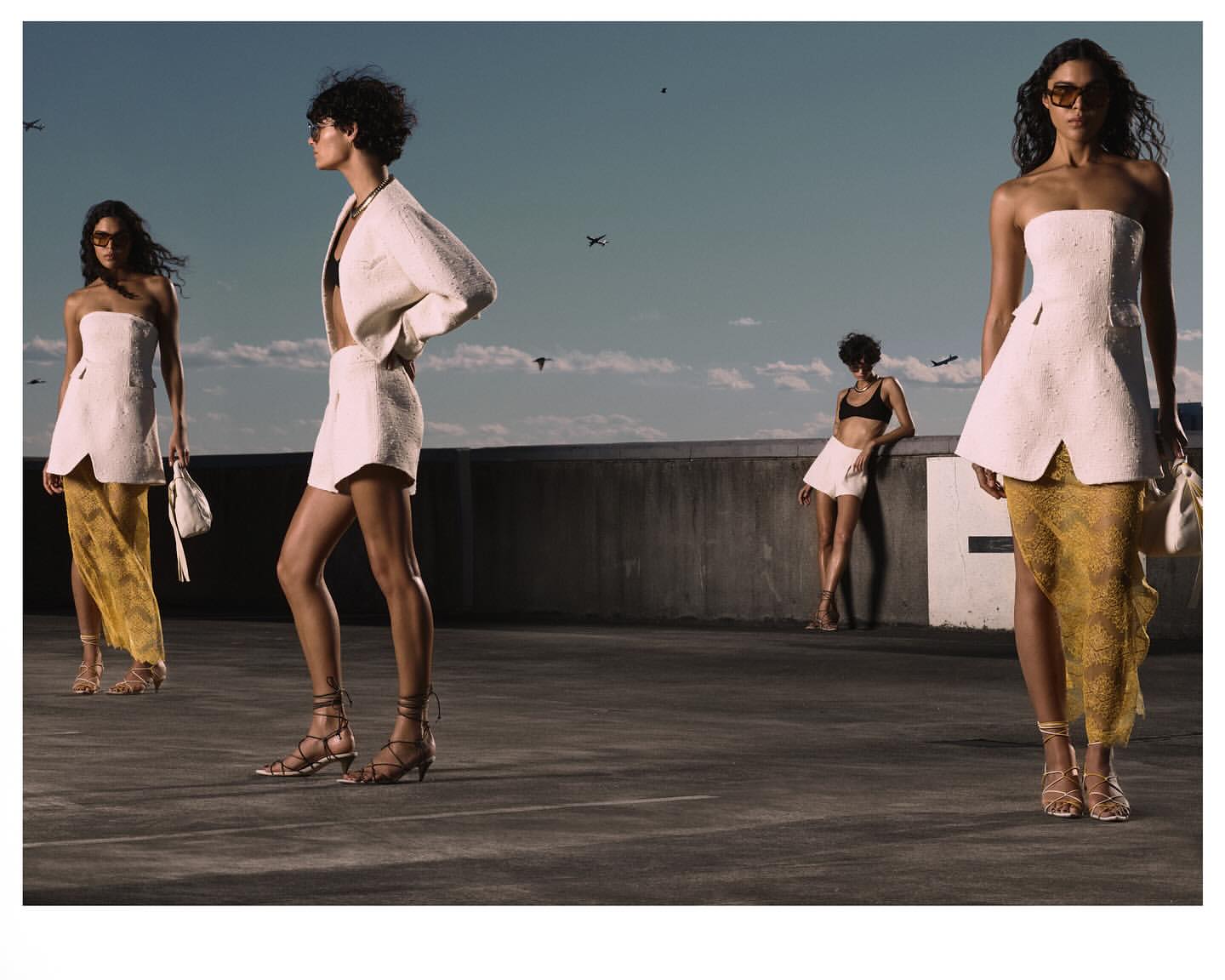 Fashion editorial shot outdoors with four young adult women wearing stylish summer clothing, including dresses, sandals, and sunglasses.