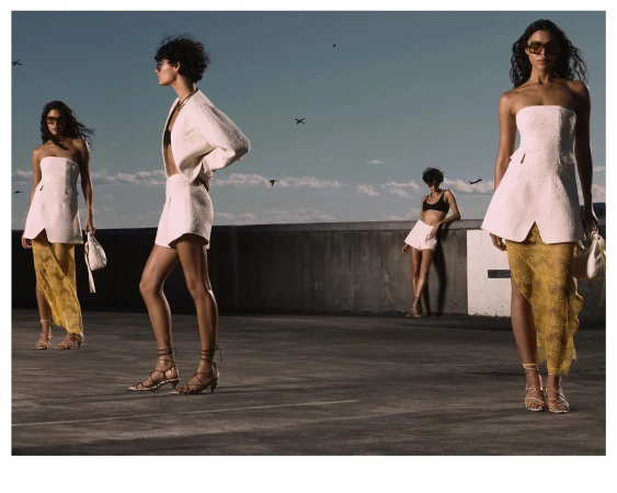 Fashion editorial shot outdoors with four young adult women wearing stylish summer clothing, including dresses, sandals, and sunglasses.