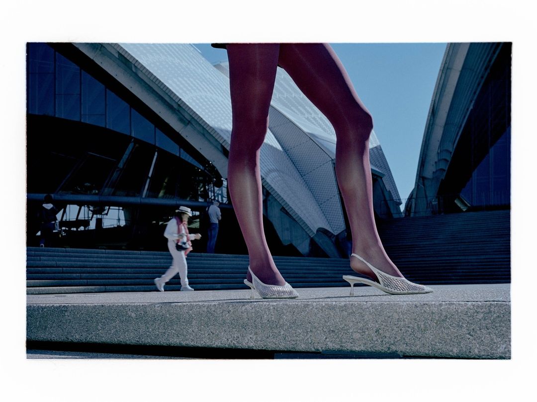 Women's legs in net slingback heels, standing on a concrete platform in front of the Sydney Opera House, captured in an outdoor shot.