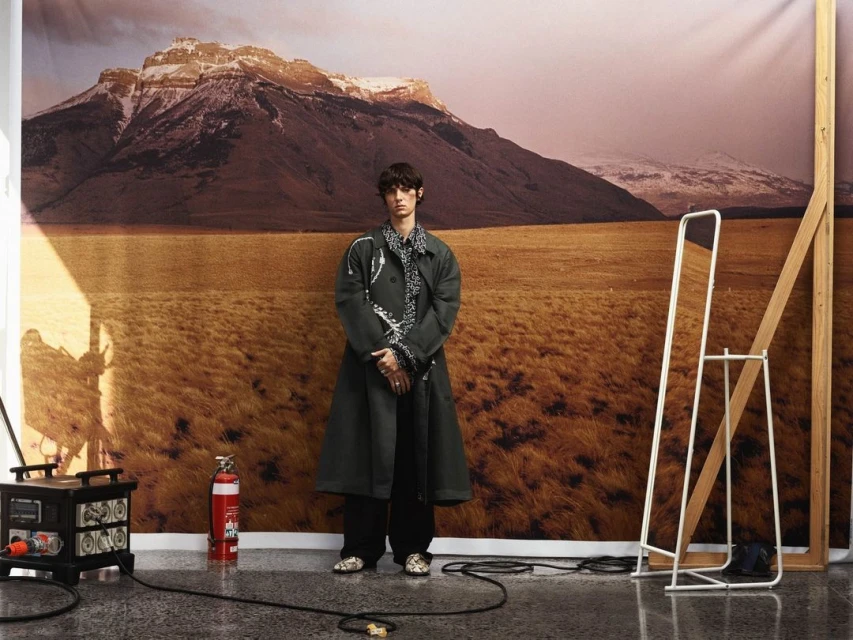 Adult male in dark coat standing in front of field and mountain backdrop inside a photo studio with camera equipment nearby.