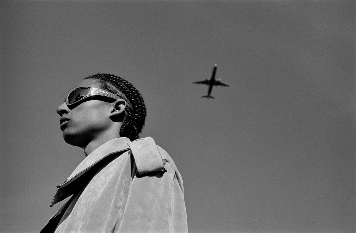 A stylish young man with braided hair and futuristic sunglasses gazes up at an airplane against a clear sky in a black and white photograph.