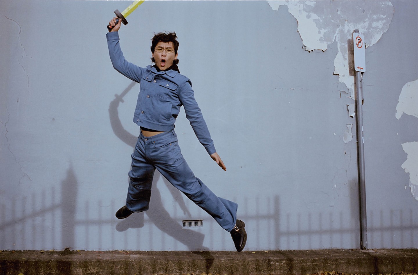 Adult is captured mid-air in a dynamic jump, holding a toy sword with an open mouth expression, wearing a denim outfit against a textured wall.