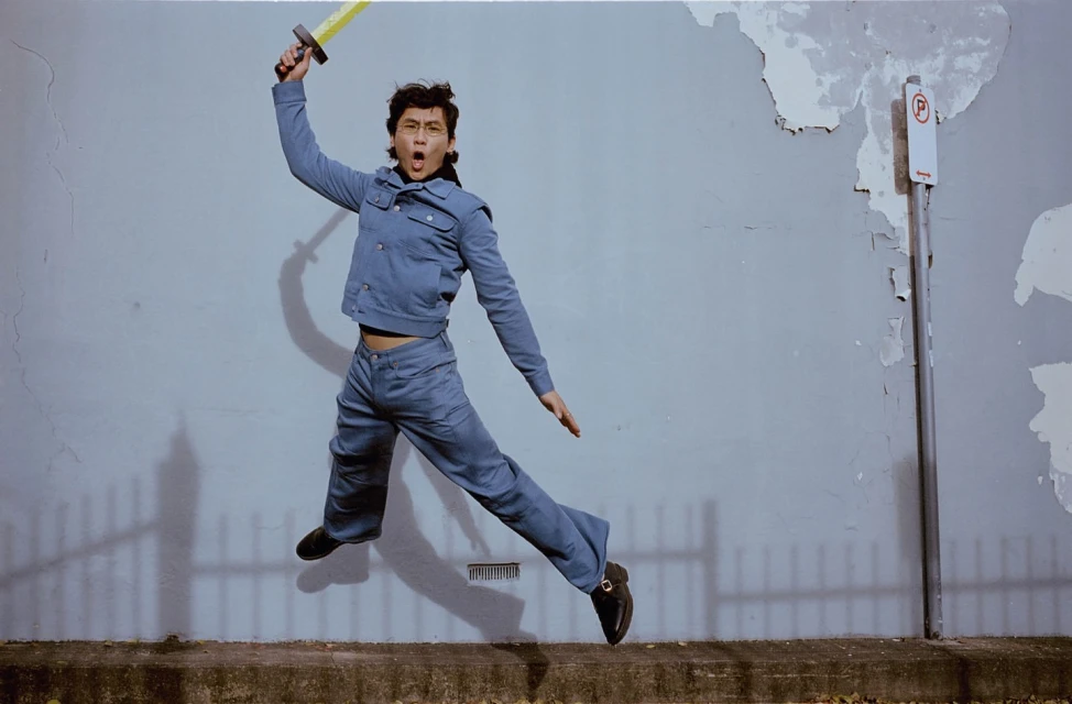 Adult is captured mid-air in a dynamic jump, holding a toy sword with an open mouth expression, wearing a denim outfit against a textured wall.