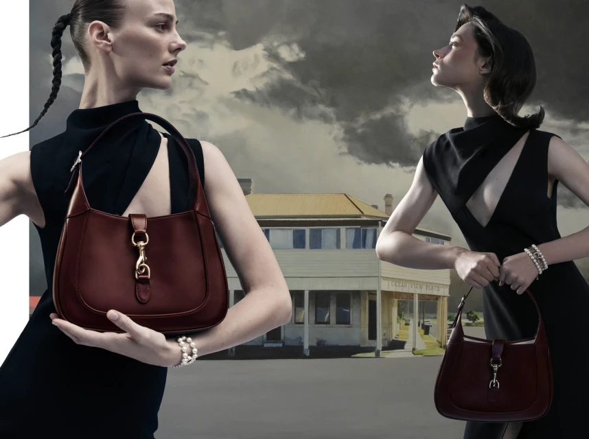 Fashion shot featuring two adult women in black dresses holding brown leather handbags with a building and cloudy sky background.