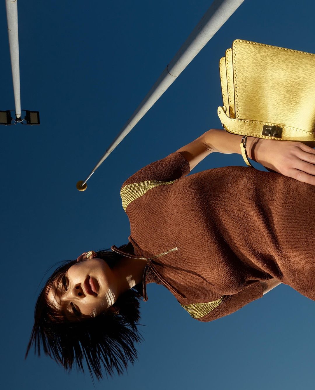 Woman in brown dress and yellow purse poses under blue sky with street lights. Fashion editorial with low angle, portrait orientation.