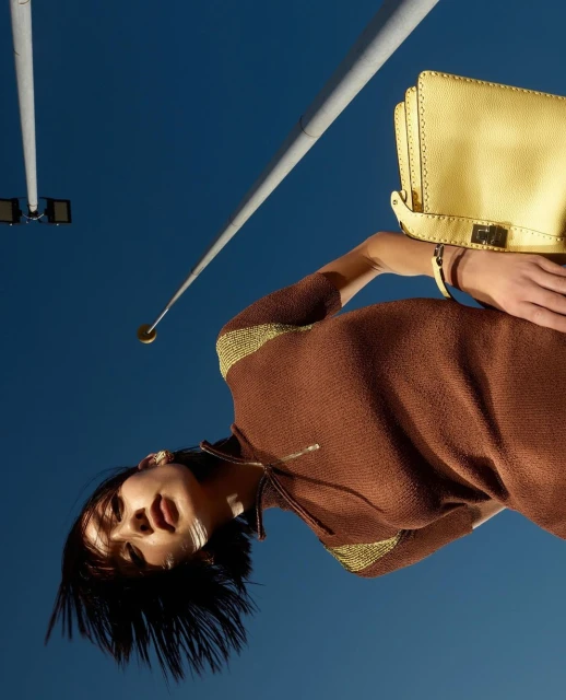 Woman in brown dress and yellow purse poses under blue sky with street lights. Fashion editorial with low angle, portrait orientation.