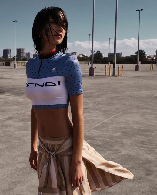 Young woman in blue and white crop top and striped skirt posing in outdoor parking lot setting on sunny day with blue sky and city view.